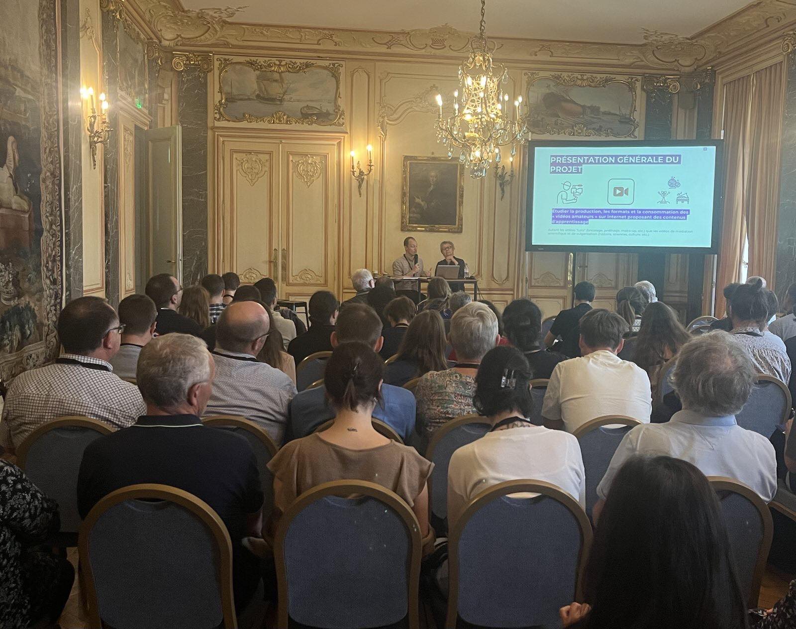 NEC 2023 - photo de la masterclass de Dominique Pasquier et Quentin Gilliotte prise du fond de la salle où l'on voit le public au premier plan et les deux intervenants et leur présentation en arrière-plan